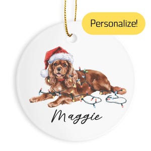 May include: A white, round Christmas ornament featuring a Cavalier King Charles Spaniel wearing a Santa hat and string lights. The dog's name, "Maggie", is written in black script below. The text "Personalize!" is in a yellow rectangle.