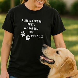May include: Black t-shirt with white text: "PUBLIC ACCESS TEST? WE PASSED THE POP QUIZ" and paw prints. A golden retriever dog is in the foreground. The shirt is worn by a person.