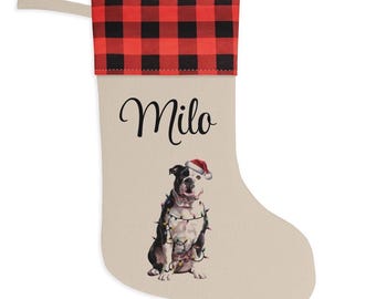 May include: A beige Christmas stocking featuring a red and black buffalo plaid cuff and a hanging loop. The stocking is personalised with the name Milo in black script and displays a graphic of a bulldog wearing a Santa hat and string lights.