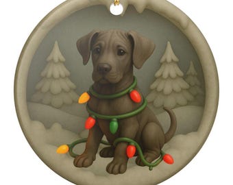 Peut inclure: Une décoration ronde en céramique représentant un chiot brun assis dans un paysage enneigé avec des sapins. Le chiot est entouré d'une guirlande verte avec des lumières orange et rouges. Un cordon doré est fixé au sommet pour suspendre.