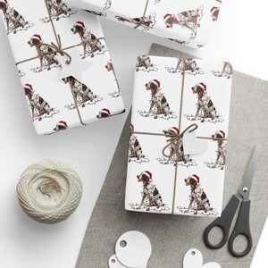 May include: White wrapping paper with a repeating pattern of a brown and white dog wearing a Santa hat and Christmas lights. The wrapped gifts are tied with twine and a heart-shaped tag. A ball of twine, scissors, and blank tags are also visible.