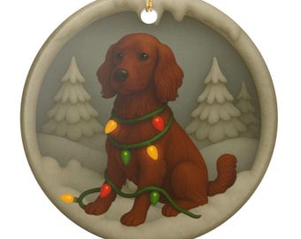 May include: A round ceramic ornament featuring a brown Irish Setter dog sitting in a snowy landscape with pine trees. The dog is wrapped in a string of colorful Christmas lights with red, yellow, and green bulbs. A gold string is attached at the top.