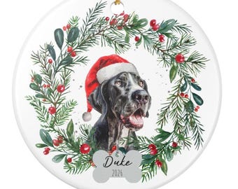 Peut inclure: Une décoration ronde en céramique blanche ornée d'une illustration aquarelle d'un chien noir portant un bonnet de Père Noël rouge, encadré par une couronne de pin vert avec des baies rouges. Une étiquette en forme d'os indique Duke et 2026.