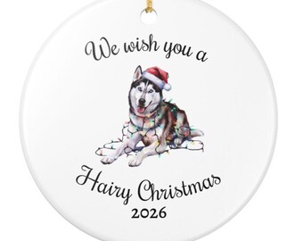 May include: A round white ceramic ornament featuring a black and white husky dog wearing a red Santa hat and tangled in colorful Christmas lights. The text reads We wish you a Hairy Christmas 2026 in black script and block lettering.