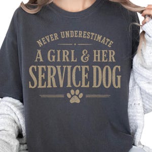 May include: A dark charcoal gray short-sleeve t-shirt featuring the gold-colored text NEVER UNDERESTIMATE A GIRL & HER SERVICE DOG printed on the front, accompanied by a small gold paw print graphic centered below the text.