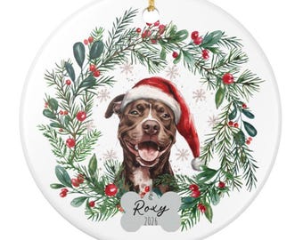 May include: A round white ceramic ornament featuring a brown dog wearing a red Santa hat, centered within a watercolor wreath of green pine branches and red berries. A bone-shaped tag hangs below the dog with the text Roxy and 2026.