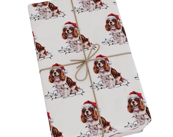 May include: A rectangular gift wrapped in white paper featuring a repeating pattern of Cavalier King Charles Spaniel dogs wearing red Santa hats and tangled in colorful Christmas string lights, tied with a thin brown twine string.