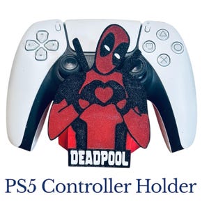 May include: A white PS5 controller with a red and black Deadpool-themed holder. The holder features a Deadpool figure with a heart-shaped design and the word "DEADPOOL" in white text. The text "PS5 Controller Holder" is below.