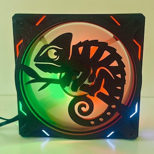May include: A black computer fan with a silhouette of a chameleon perched on a branch. The fan has a colorful LED light display with green, orange, and blue hues. The fan is square-shaped.
