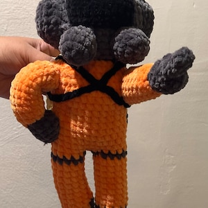 May include: A crocheted plush toy in the shape of a person wearing an orange jumpsuit with black accents and a black crocheted head.