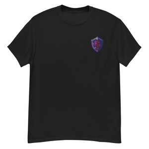 May include: A black short-sleeve t-shirt featuring a small pixel art graphic on the upper left chest. The graphic depicts a purple and grey shield with a red emblem in the center, rendered in a distinct retro video game aesthetic style.