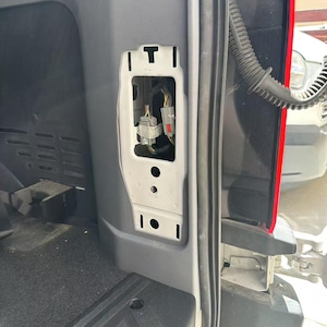 May include: A white plastic bracket with a T-shaped top and rectangular cutouts, mounted inside a vehicle's interior. The bracket houses electrical connectors with colored wires. The vehicle's interior is gray and black.