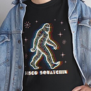 May include: A black t-shirt featuring a graphic of a walking Bigfoot with a multi-colored chromatic aberration effect. The design includes starburst shapes and the text DISCO SQUATCHIN in a retro font with a similar layered color effect.