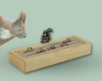 May include: A wooden ring holder with a light brown finish and a beige velvet interior. The holder has five slots for rings and a silver squirrel figurine in the center.