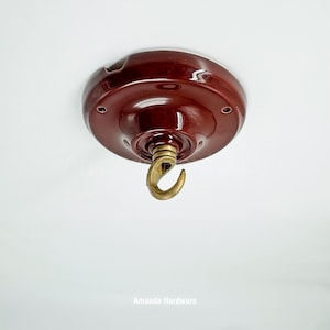 May include: A maroon ceiling canopy with a brass hook. The canopy is round with a glossy finish and has several small holes. The hook is curved and appears to be made of brass. The text "Amanda Hardware" is at the bottom.
