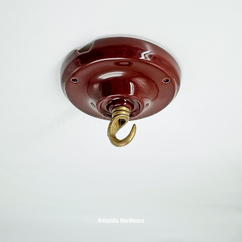 May include: A maroon ceiling canopy with a brass hook. The canopy is round with a glossy finish and has several small holes. The hook is curved and appears to be made of brass. The text "Amanda Hardware" is at the bottom.