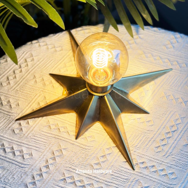 May include: A brass-colored metal star-shaped light fixture with a central circular glass bulb emitting a warm glow. The fixture features multiple pointed rays extending from the center and includes the text Amanda Hardware printed on the surface.
