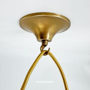 May include: A close-up of a brass-coloured ceiling canopy with a matching hook and a gold-coloured fabric-covered electrical cord. The text "Amanda Hardware" is visible at the bottom of the image. The fixture is designed for hanging a light.