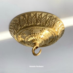 May include: Gold ceiling medallion with a decorative leaf design and a hook for hanging a chandelier. The medallion is 15 cm in diameter.