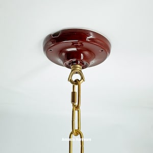 Puede incluir: Primer plano de una roseta de techo de color rojo oscuro con una cadena de latón. La roseta es redonda con una ligera forma de cúpula y varios agujeros pequeños. La cadena es de metal dorado.