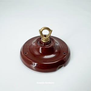 May include: A maroon ceiling canopy with a brass loop and threaded post. The canopy has a glossy finish and two small holes. The text "Amanda Hardware" is printed on the bottom.