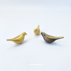 May include: Three small bird figurines, two are gold and one is a darker bronze color. The birds are arranged in a triangular formation on a white background.