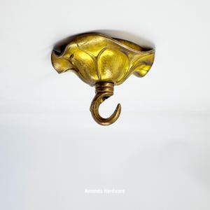May include: A gold-coloured ceiling hook with a decorative, leaf-like canopy. The hook is curved and hangs from a detailed, ornate base. The fixture is mounted on a white surface.
