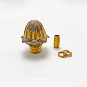 May include: A gold-toned metal finial with a fluted design and a decorative base. It comes with a threaded metal rod and two metal rings.