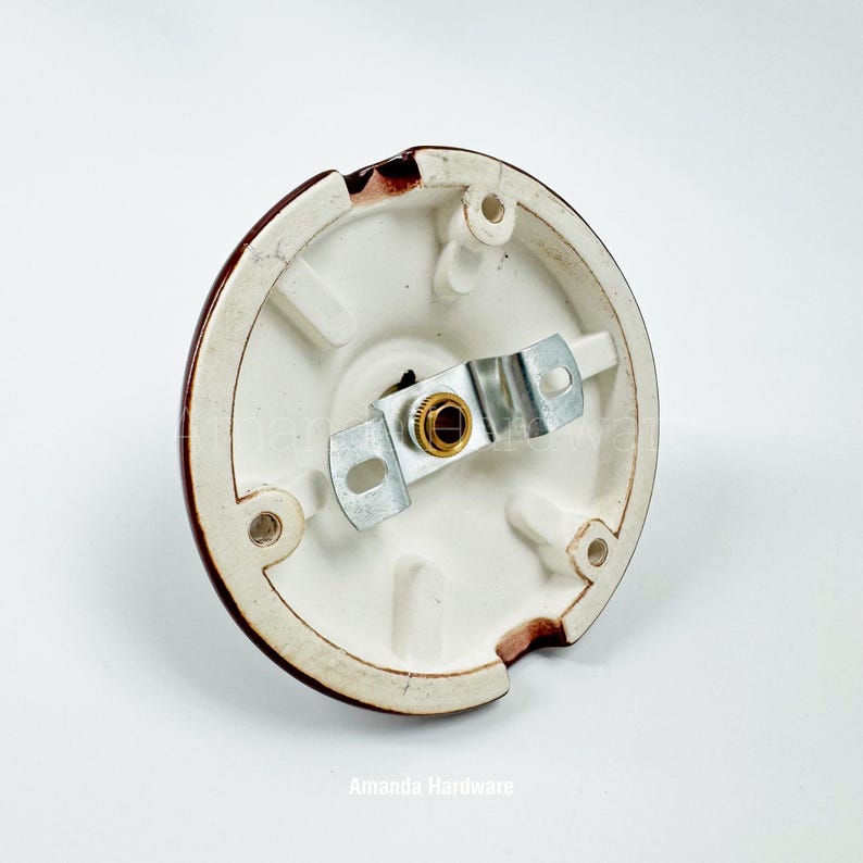 May include: A white ceramic light fixture base with a brown rim. The base has a metal bracket and a gold-colored center. The fixture has three screw holes and a small notch. The text "Amanda Hardware" is at the bottom.