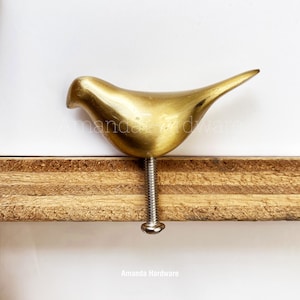 May include: A gold bird-shaped cabinet knob with a silver screw. The knob is mounted on a piece of wood.