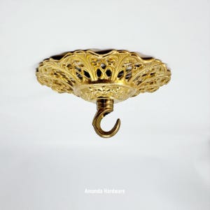 May include: Ornate gold-colored ceiling canopy with a decorative filigree design. Features a central hook for hanging a light fixture or other decorative items. The canopy is round and has a detailed, vintage aesthetic.