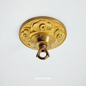 May include: A gold-colored ceiling canopy with a floral design. The canopy has a circular shape with embossed floral patterns and a central loop for hanging. The text "Amanda Hardware" is at the bottom.