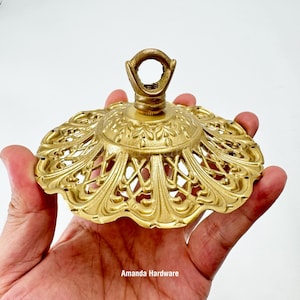 May include: Ornate, gold-colored ceiling canopy with a decorative, openwork design. The canopy has a central loop for hanging and is likely used for lighting fixtures. The text "Amanda Hardware" is visible.