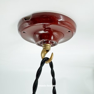 May include: A maroon ceiling canopy with a brass hook and a black twisted cord. The canopy has a white rim and several small holes. The hook is attached to the canopy's center. The cord hangs down from the hook.