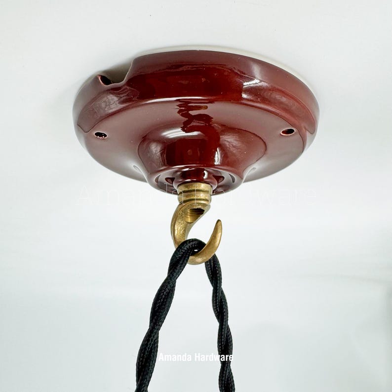 May include: A maroon ceiling canopy with a brass hook and a black twisted cord. The canopy has a white rim and several small holes. The hook is attached to the canopy's center. The cord hangs down from the hook.