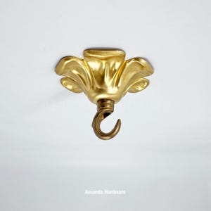 May include: A gold-coloured ceiling hook with a floral design. The hook is curved and hangs from a decorative, flower-shaped base. The base has a textured, metallic finish. The text "Amanda Hardware" is at the bottom.