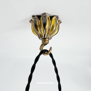 Peut inclure: Un luminaire de plafond décoratif avec un motif floral dans des tons or et argent. Un cordon noir torsadé est suspendu à un crochet en laiton sous le luminaire. Le texte "Amanda Hardware" est visible en bas.