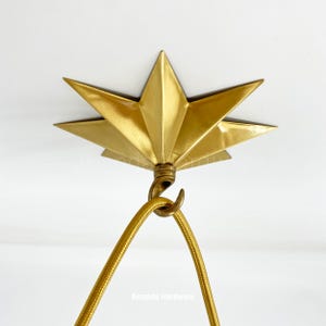 May include: A gold star-shaped ceiling plate with a hook for hanging a light fixture. The plate is attached to a white ceiling. The text "Amanda's Hardware" is visible below the plate.