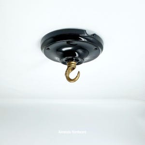 May include: A black ceiling hook with a brass hook. The ceiling rose is round and has a glossy finish. The hook is designed for hanging light fixtures or other decorative items. The text "Amanda Hardware" is at the bottom.