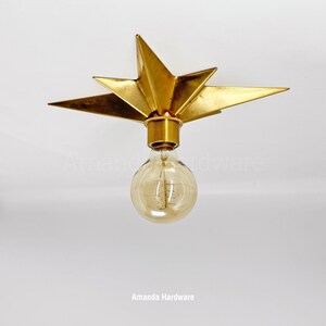 May include: A gold-colored metal star-shaped flush mount light fixture featuring a central cylindrical socket and a round, clear glass Edison-style light bulb with a visible filament. The fixture has multiple pointed rays extending outward.