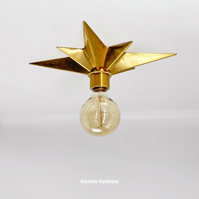 May include: A gold-colored metal star-shaped flush mount light fixture featuring a central cylindrical socket and a round, clear glass Edison-style light bulb with a visible filament. The fixture has multiple pointed rays extending outward.