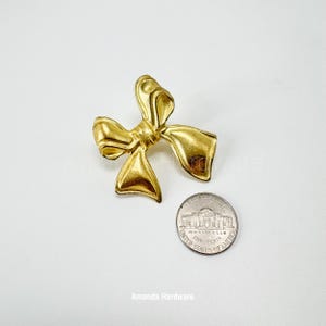 Może przedstawiać: Złota metalowa kokardka o lekko fakturowanej powierzchni. Kokardka jest mniej więcej wielkości polskiej monety pięciogroszowej.