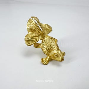 Peut inclure: Une figurine décorative en forme de poisson doré, avec des écailles détaillées et des nageoires fluides. Le poisson est positionné en biais, mettant en valeur sa surface texturée et son design élégant. La figurine est probablement en métal.