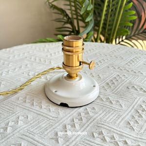 May include: A vintage-style lamp holder with a white ceramic base and a brass-coloured socket. A gold-coloured twisted wire extends from the base. The text "Amanda Hardware" is visible on the base. This is a lighting component.