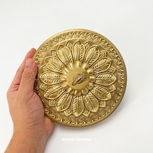 May include: A gold-coloured, circular decorative hardware piece with an ornate floral design. It features a central keyhole and is likely made of metal. The text "Amanda Hardware" is visible at the bottom.