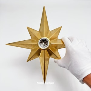 May include: A gold-colored, eight-pointed star-shaped light fixture component with a central circular socket. The metallic surface features a smooth finish with subtle variations in tone. The object is symmetrical with sharp, elongated points.