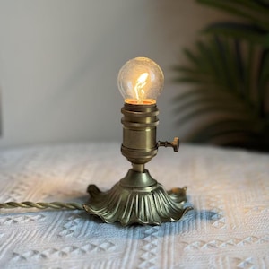 May include: A vintage brass lamp with a decorative base and a small, round light bulb. The lamp is lit, emitting a warm glow. The cord is a twisted gold colour. The lamp is on a white patterned surface.