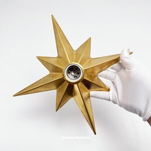 May include: A gold-colored star-shaped light fixture with multiple pointed rays extending from a central circular socket. The metallic surface features a smooth finish with subtle variations in tone. The text Amanda Hardware is visible.