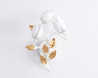 May include: White metal wall sconce featuring a floral design with three tulip-shaped lights and gold-colored leaves. The sconce is mounted on a white surface. The text "Amanda Lighting" is visible.