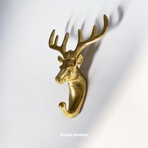 May include: Gold deer head wall hook. The hook is shaped like a deer head with antlers and a curved hook at the bottom. The hook is mounted on a white wall. The text "Amanda Hardware" is printed below the hook.
